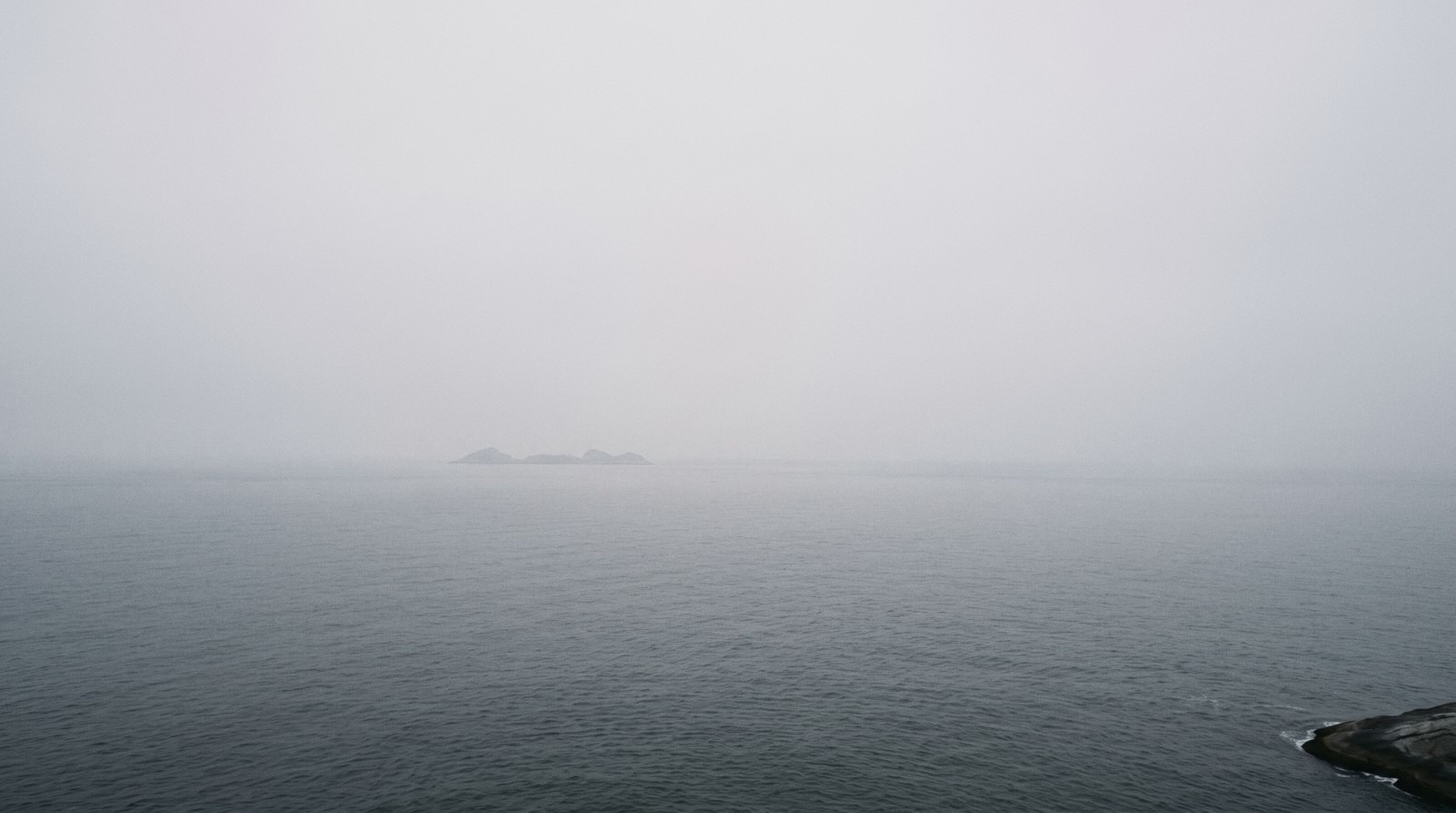 The Cagarras islands in morning fog off Rio, seen from Leblon. A flat grey sea and a dark rock at the edge.