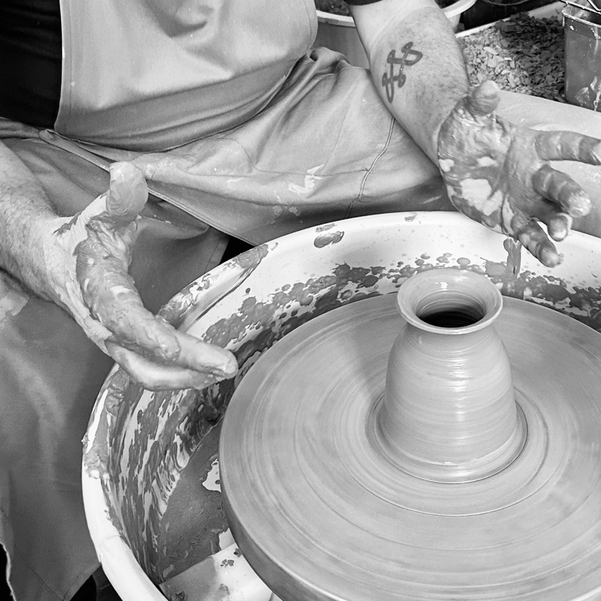 Clay-covered hands open beside a small vase turning on a potter&rsquo;s wheel, in black and white.